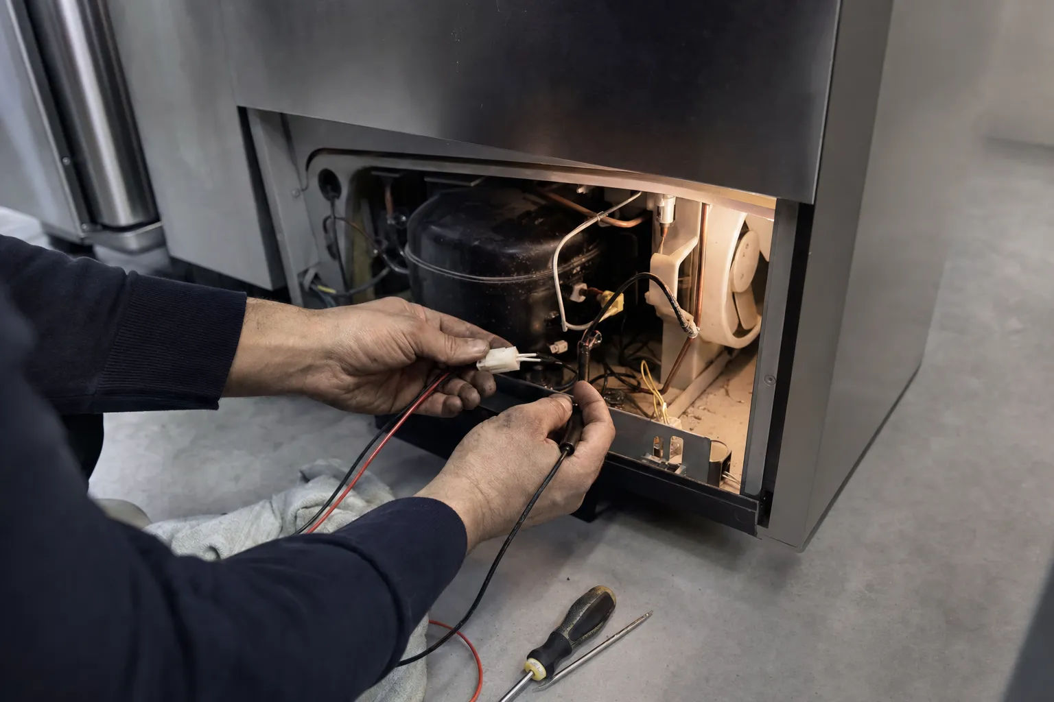 Multimeter probes on a wiring connector beside the compressor and condenser fan, lower panel removed