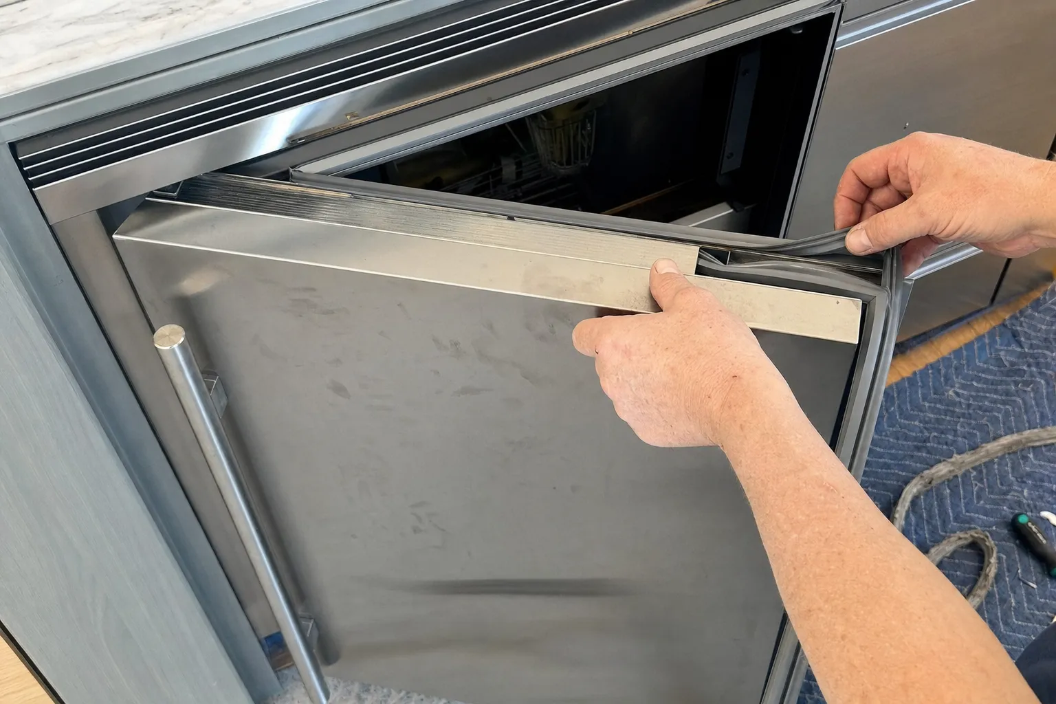 Rubber gasket peeled back at the corner of a stainless undercounter door while the other hand lifts the door trim strip