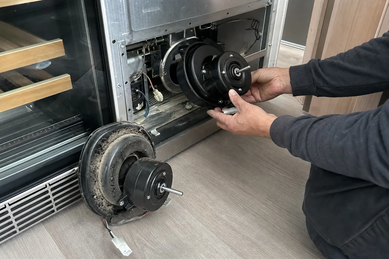 Hands fitting a new fan motor into the lower bay of a glass-door wine cabinet, the dust-caked old motor on the floor